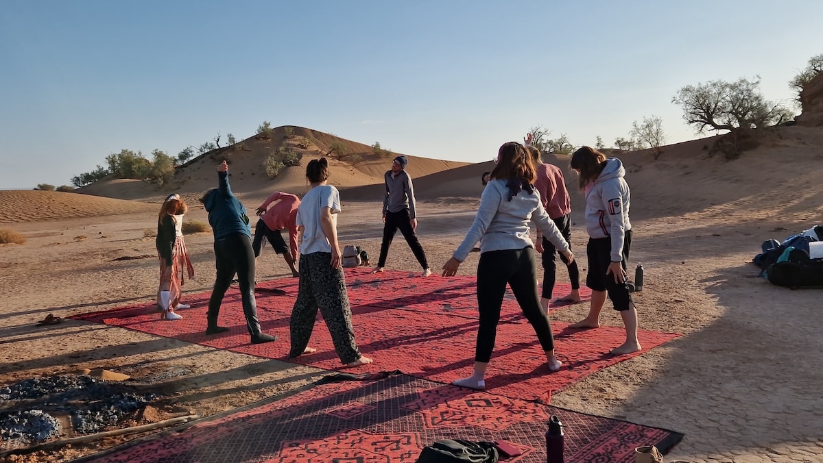 Groupe de personnes en train de réaliser des mouvements matinaux sur des tapis dans le désert, sous un ciel clair, entourés de dunes.